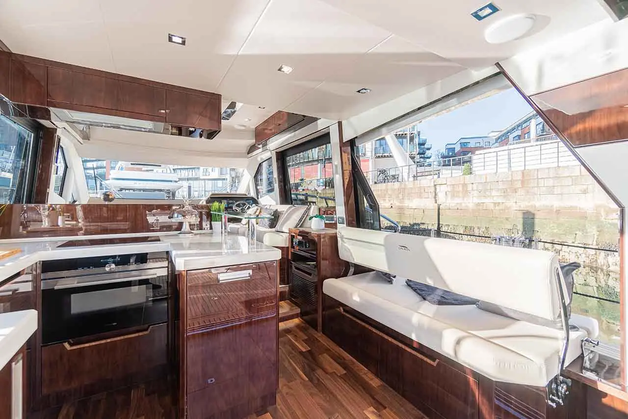 Modern boat interior featuring a sleek kitchen area, lounge seating, and large windows showing waterfront views.