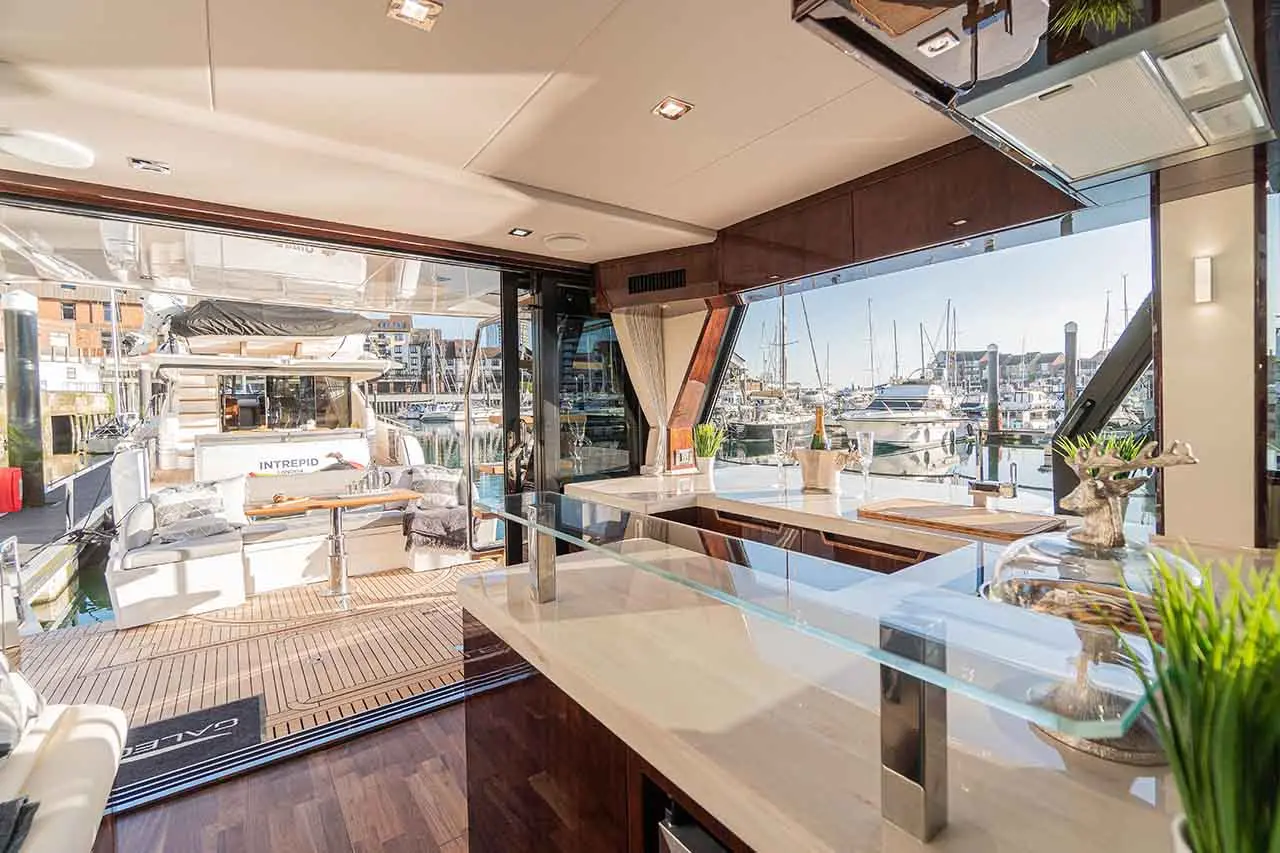 Luxury yacht interior with modern kitchen, large windows revealing a marina view, and stylish decor including plants and glassware.