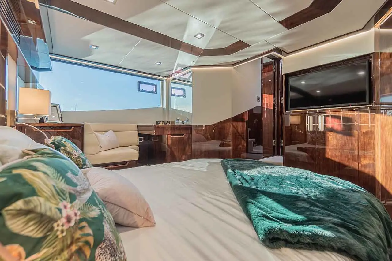 Luxurious yacht bedroom with plush bedding, green throw, sofa, and large windows showcasing a blue sky. Wall-mounted TV included.