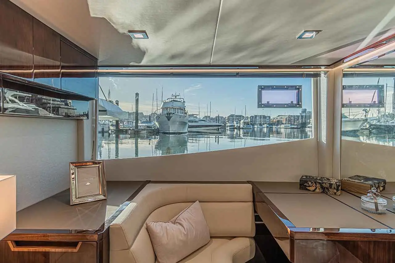 Luxurious boat interior with a cream sofa, glass windows, and a view of a tranquil marina with yachts reflecting in the water.