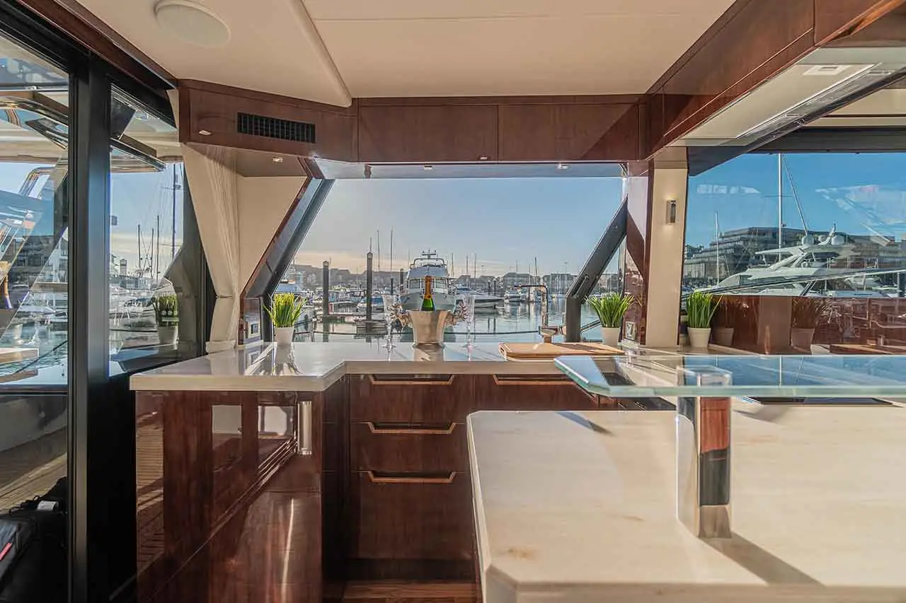 Modern yacht interior featuring a sleek kitchen, large windows overlooking a marina, and a champagne bottle in a cooler.