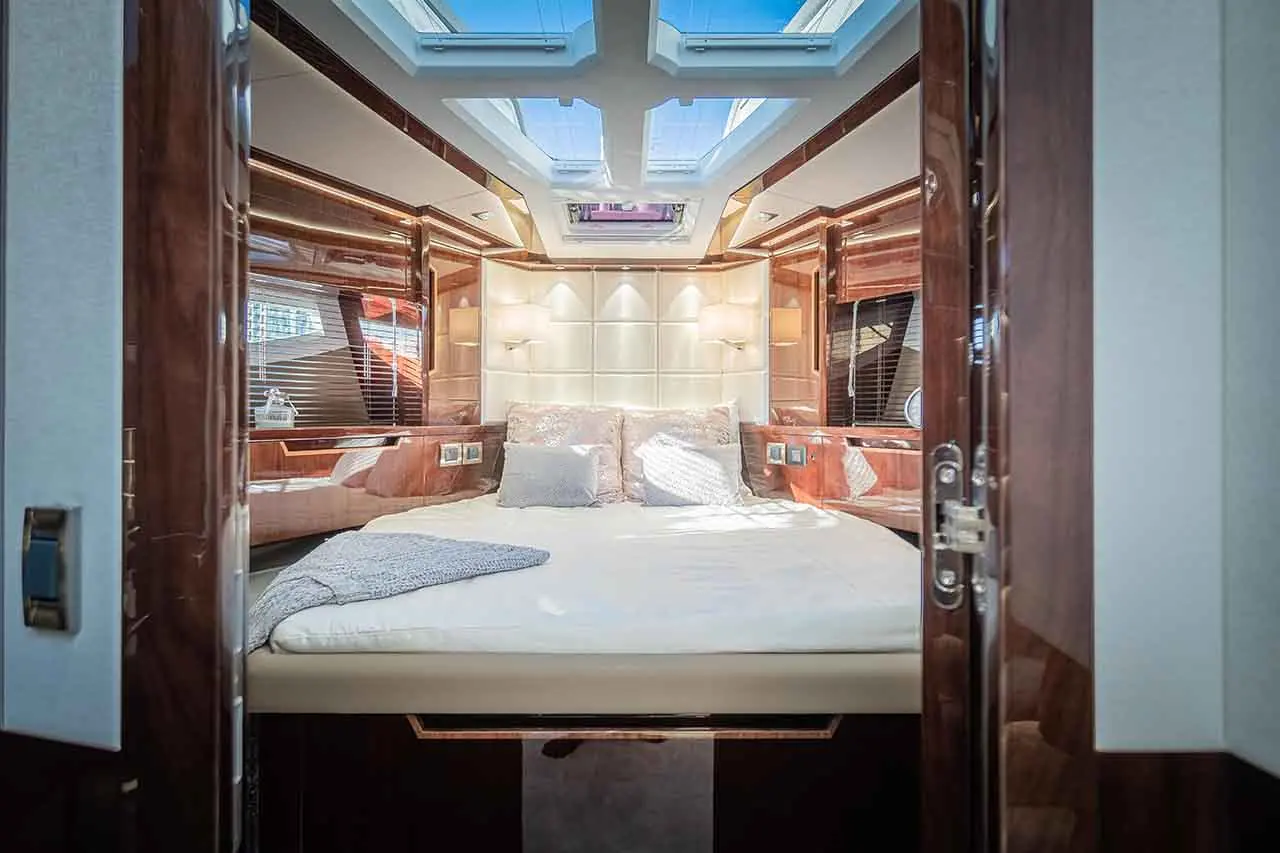 Luxurious yacht interior featuring a double bed, plush pillows, and natural light from a skylight. Elegant wooden finishes visible.