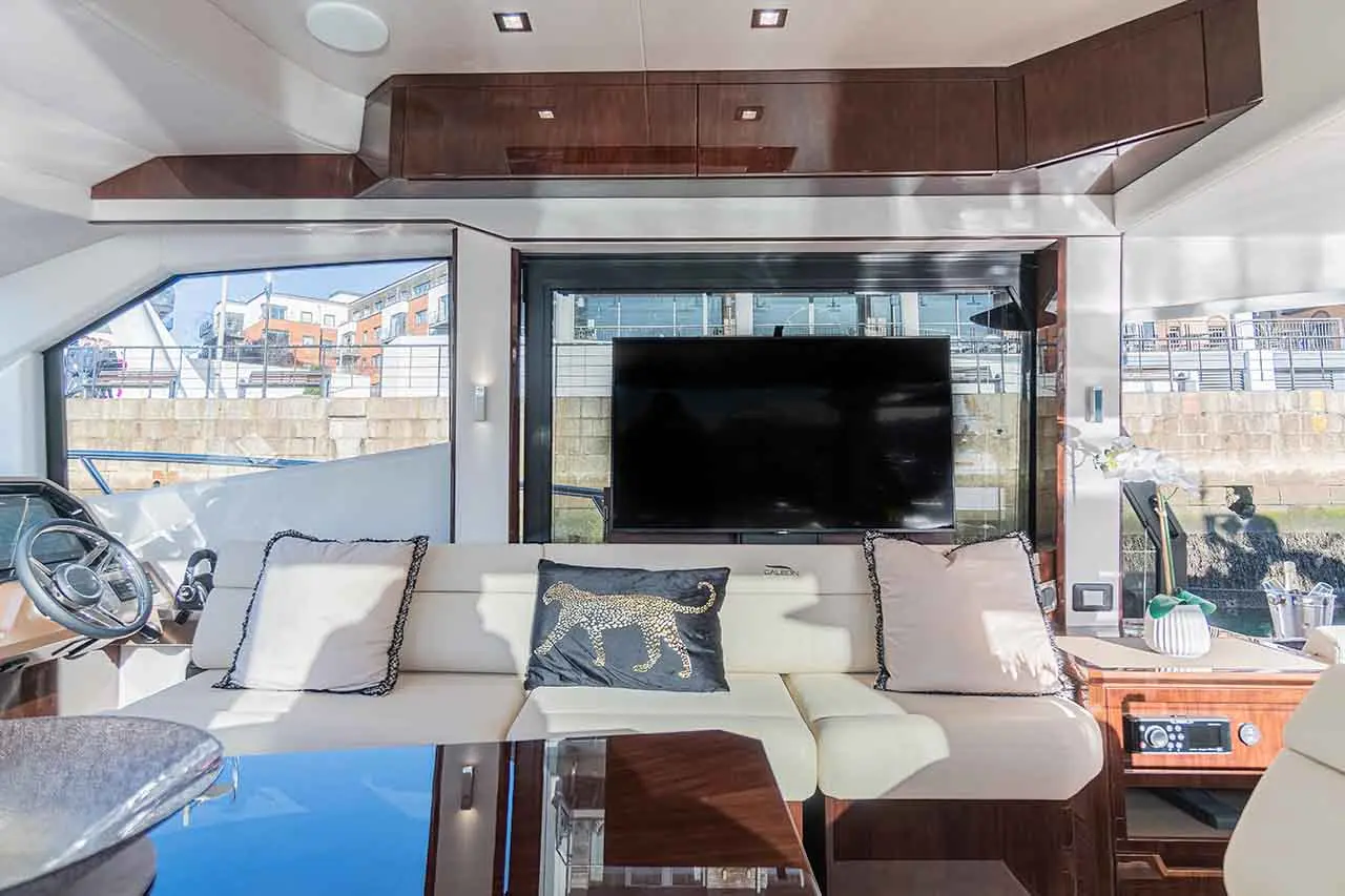 Luxurious interior of a yacht with a white sofa, decorative cushions, a large TV, and panoramic windows overlooking the marina.