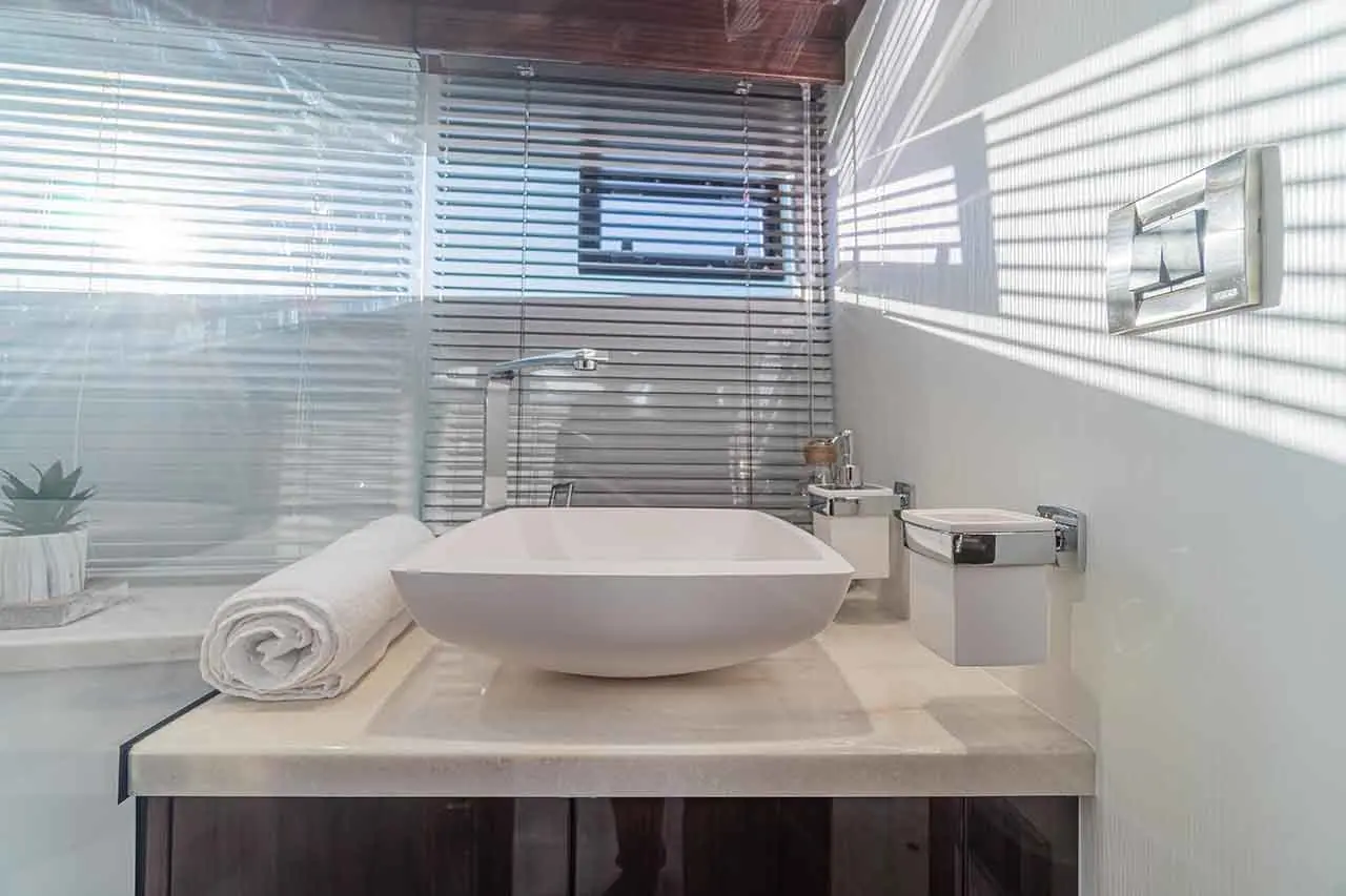Modern bathroom with a white vessel sink, rolled towel, plants, and chrome fittings, illuminated by natural light through blinds.