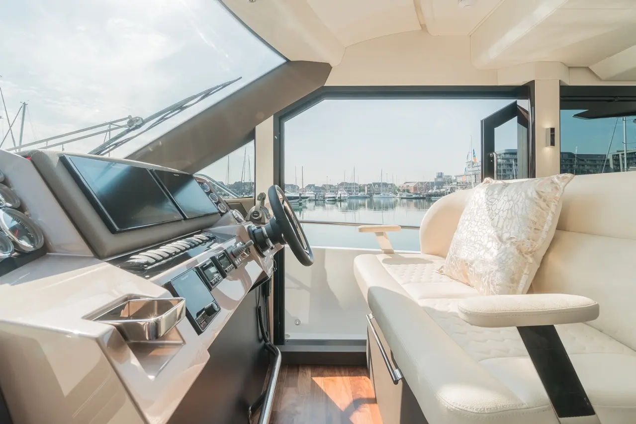 Luxury yacht interior showcasing a helm with multiple screens, steering wheel, and plush seating overlooking a marina.