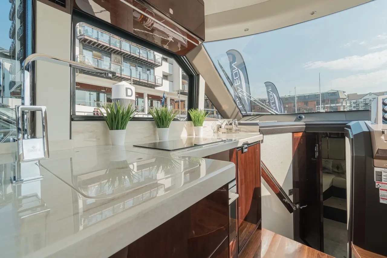 Modern yacht interior featuring a sleek kitchen area, large windows, potted plants, and views of nearby buildings.