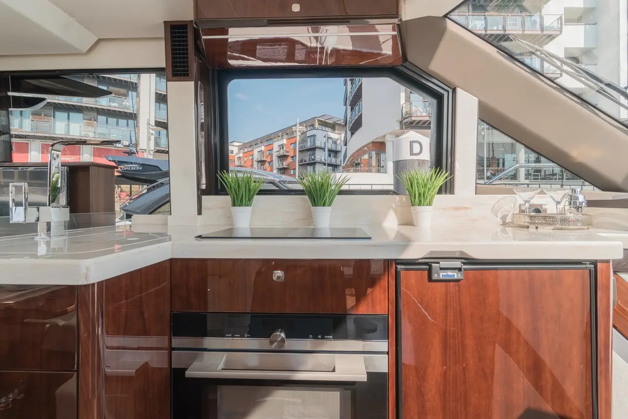 Sleek boat kitchen with glossy cabinets, potted plants, and panoramic windows showcasing modern buildings and a blue sky.