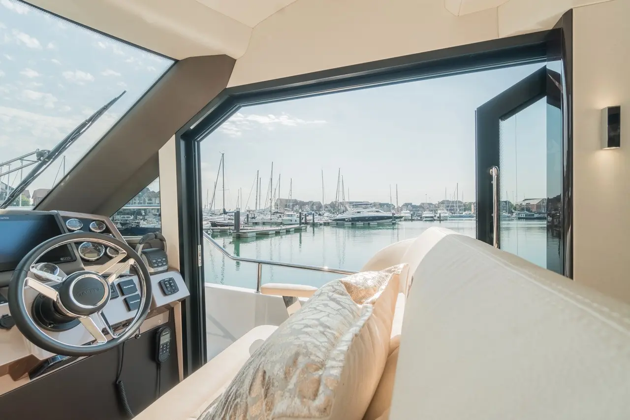 Interior of a luxury boat showing the steering wheel and soft furnishings, with a marina view through large windows.