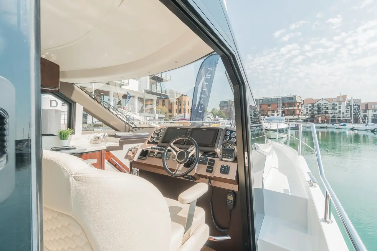 Luxury yacht cabin with a modern helm, steering wheel, and control panel, overlooking a serene marina with buildings in the background.