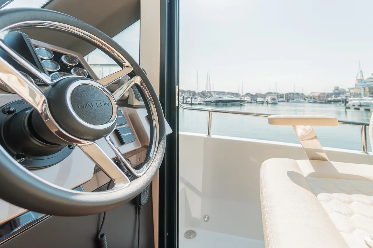 Galeon boat interior featuring a sleek steering wheel and comfortable seat, overlooking a marina with moored yachts.
