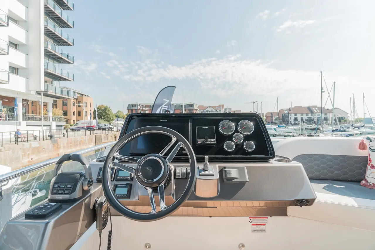 Boat cockpit featuring a steering wheel, control panel, gauges, and a view of the marina with buildings and boats.