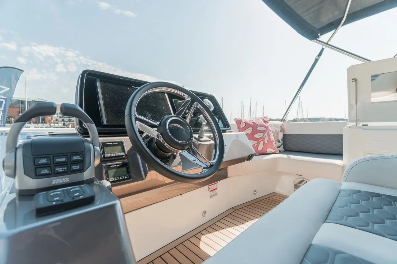 Boat interior showcasing a steering wheel, control panel, and comfortable seating, with a sunny marina view outside.