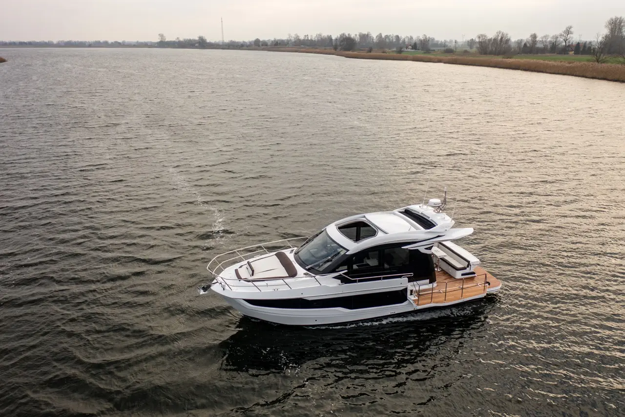 A luxury motorboat navigates a calm river with a cloudy sky, showcasing its sleek design and wooden deck.