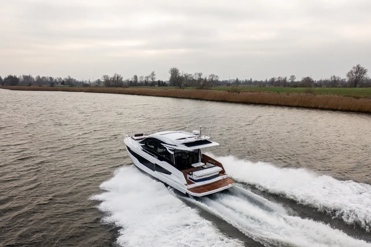 A sleek white motorboat navigates a calm river, creating waves, with lush greenery and cloudy skies in the background.