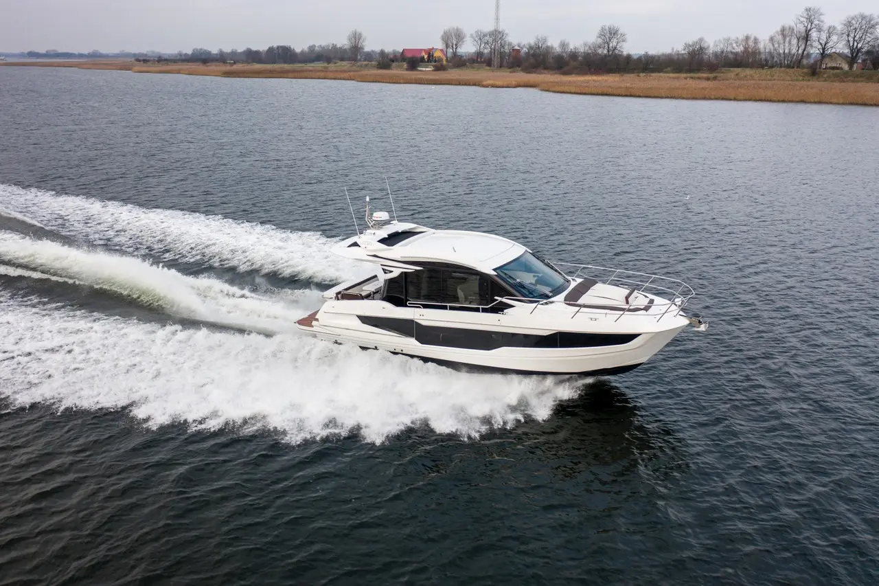 A sleek white boat speeds across a calm waterway, leaving a frothy wake, with trees and a distant building in the background.