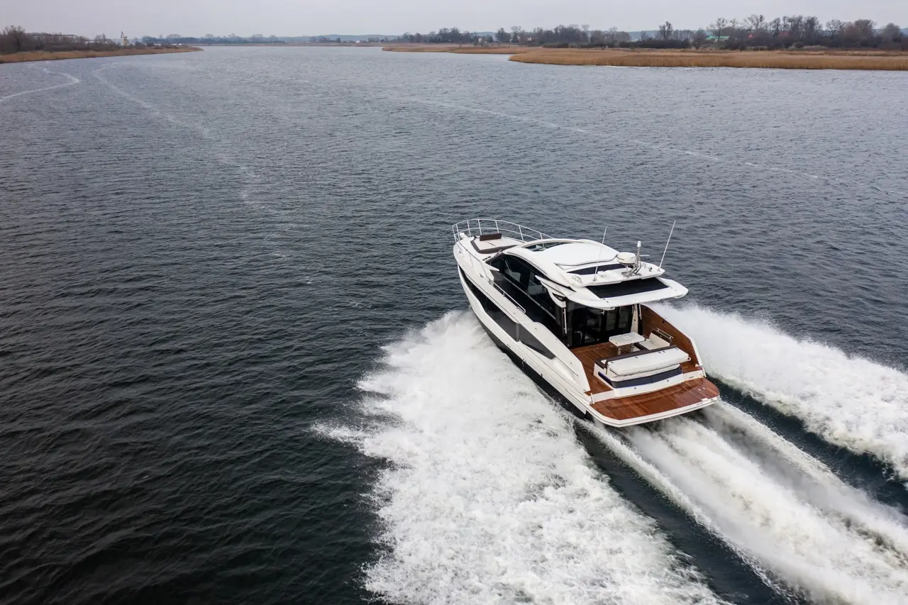 Luxury motor yacht gliding through calm water, leaving a white wake, with a scenic shoreline in the background.