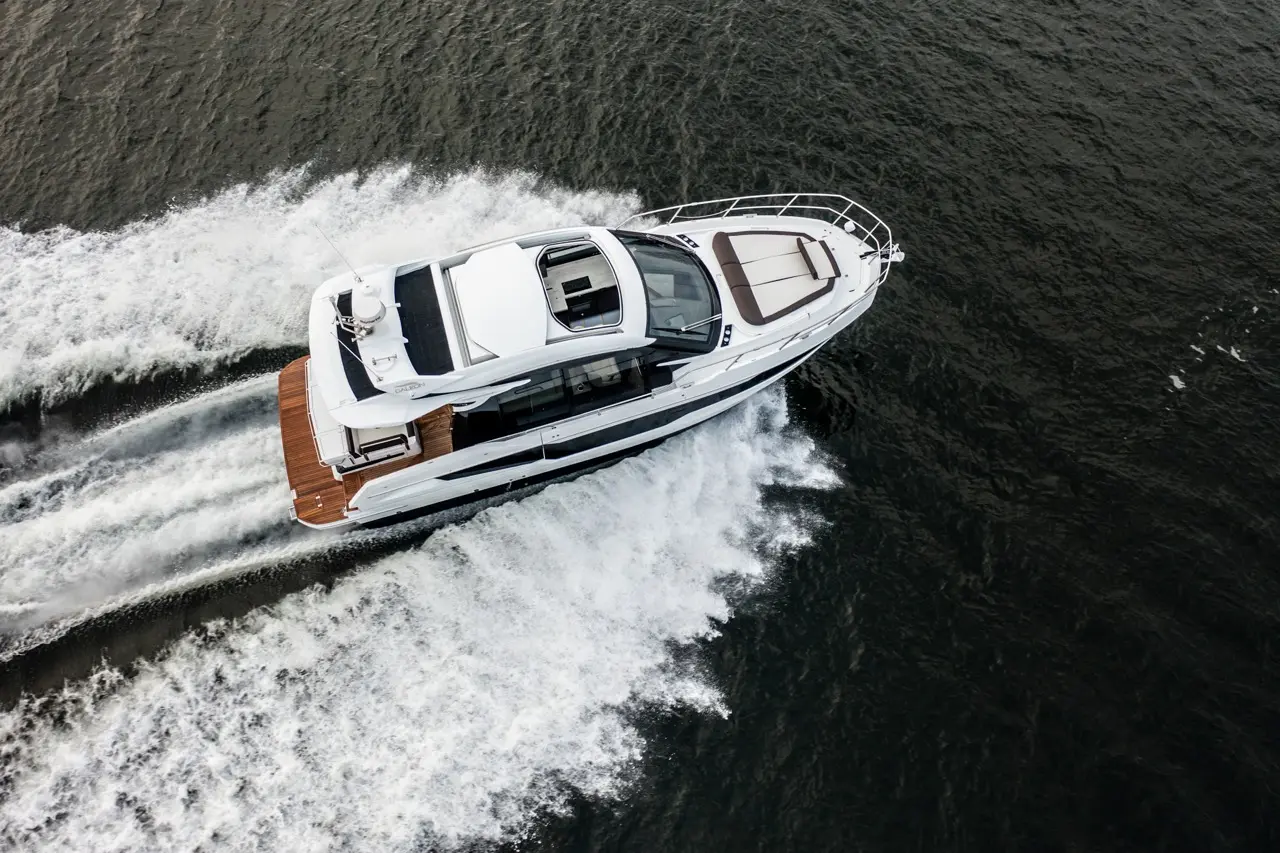 A luxury motor yacht speeds across dark water, leaving white frothy waves in its wake under an overcast sky.
