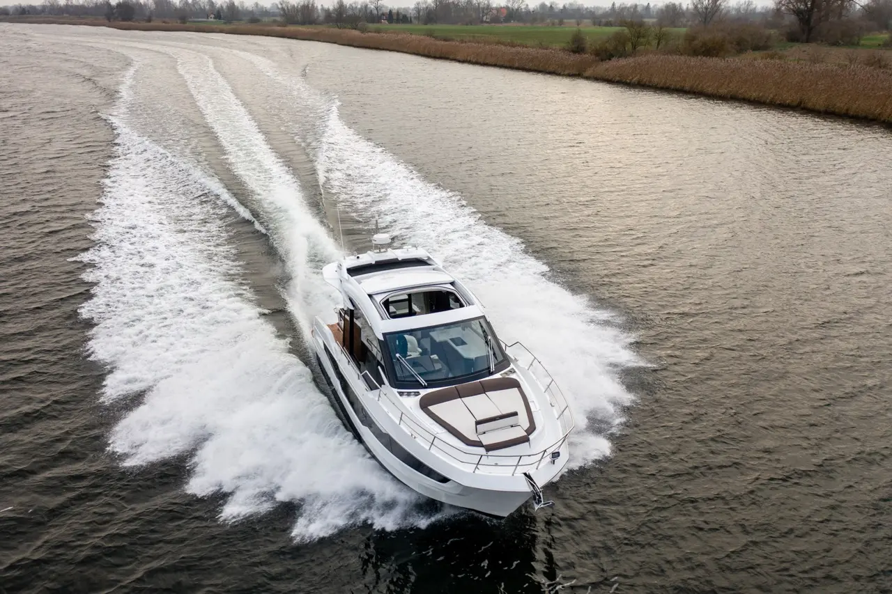 A sleek motorboat speeds through calm water, leaving a wake, surrounded by greenery and distant trees in a tranquil setting.