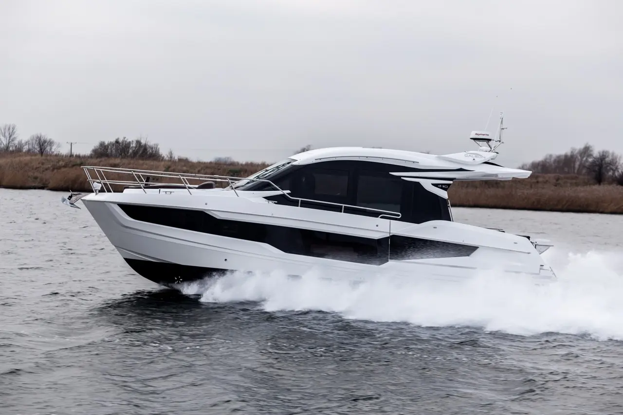 A sleek white motor yacht speeds across a calm waterway, creating splashes with its bow, set against a grey sky.