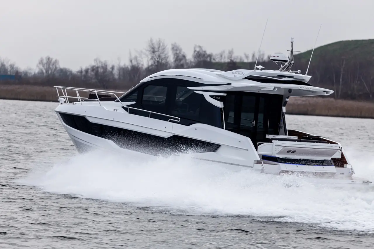 A sleek white motorboat speeds across water, creating splashes, with a blurred landscape of trees in the background.