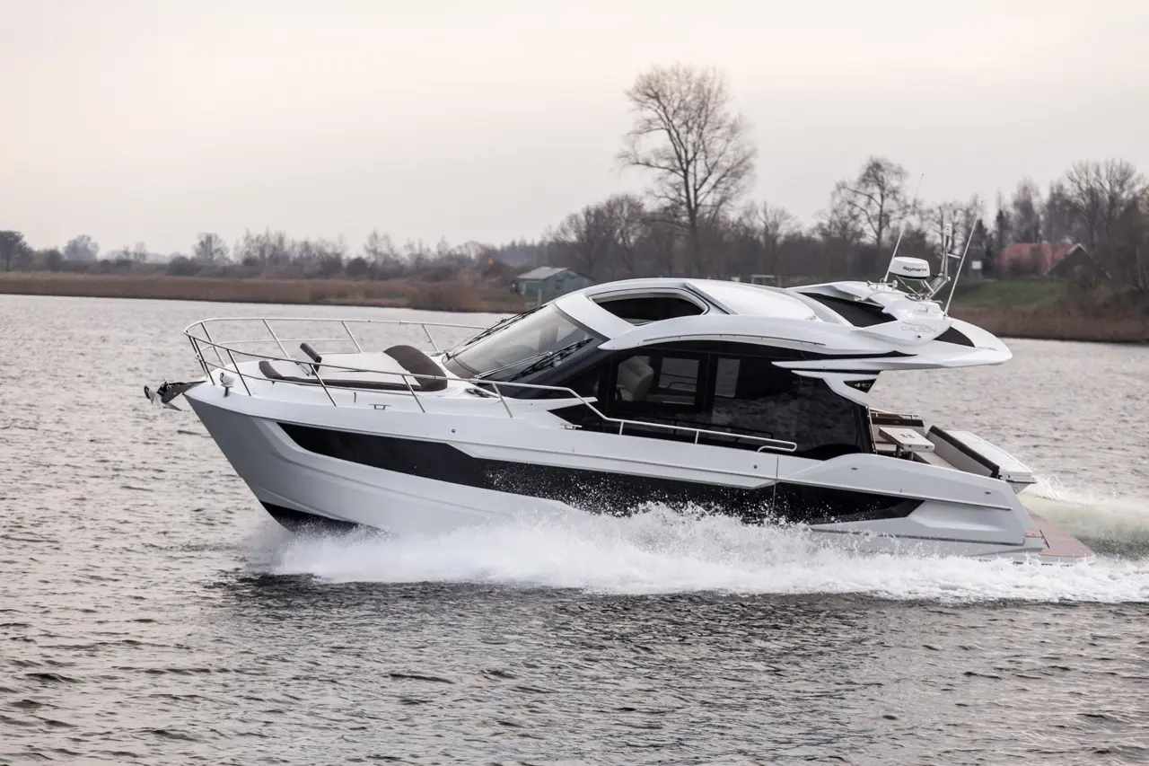 Stylish motorboat cruising on a tranquil waterway, creating waves, with trees and a distant building on the shore.