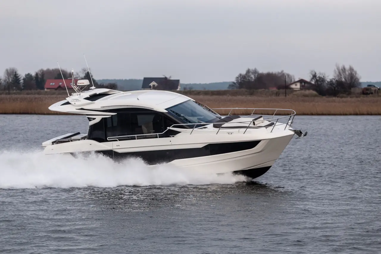 A sleek white motorboat speeds across calm waters, leaving a trail of spray, with houses and trees in the background.