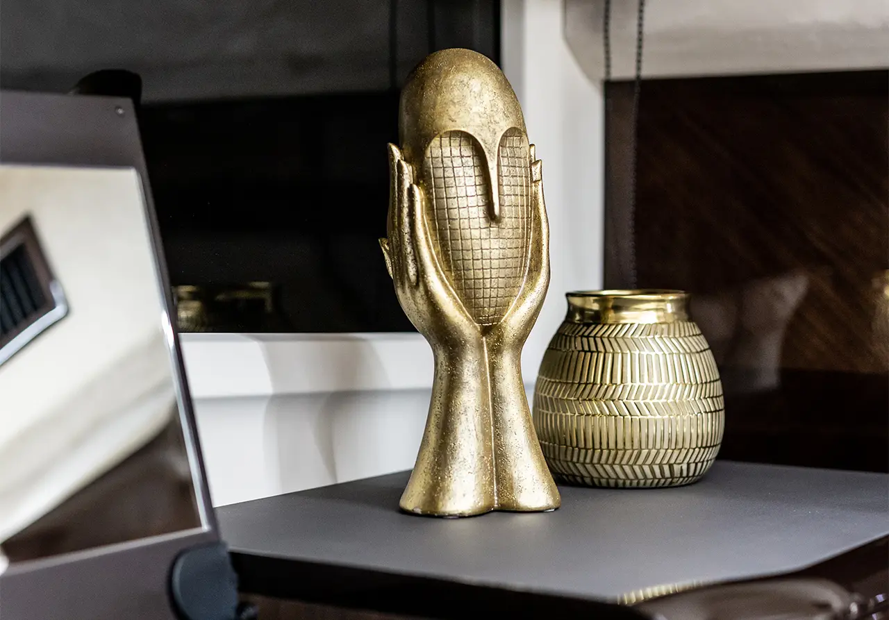 A gold sculpture of hands holding an oval figure, beside a decorative gold vase with a geometric pattern.