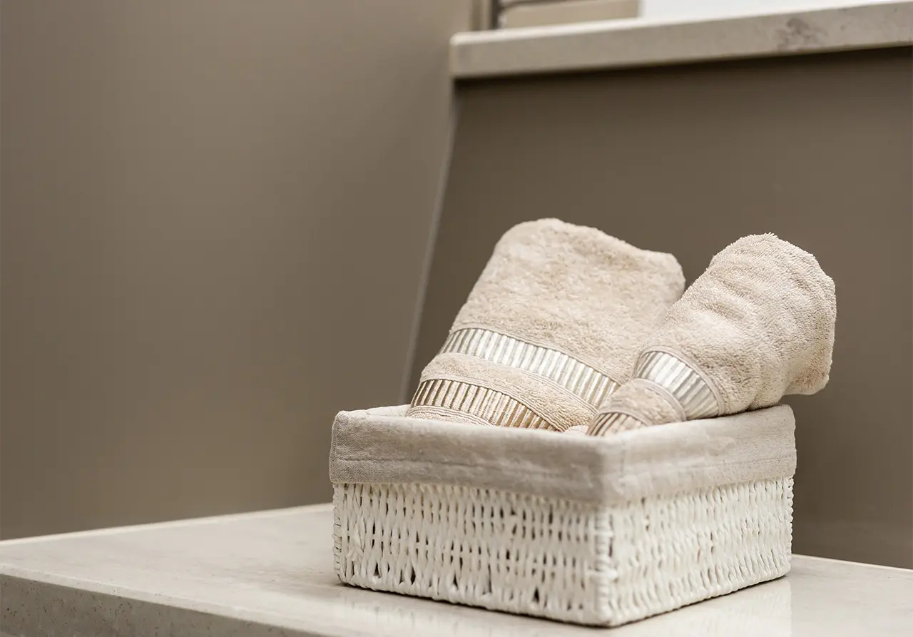 Neatly folded beige towels in a wicker basket on a light-coloured countertop, with a soft, minimalist background.