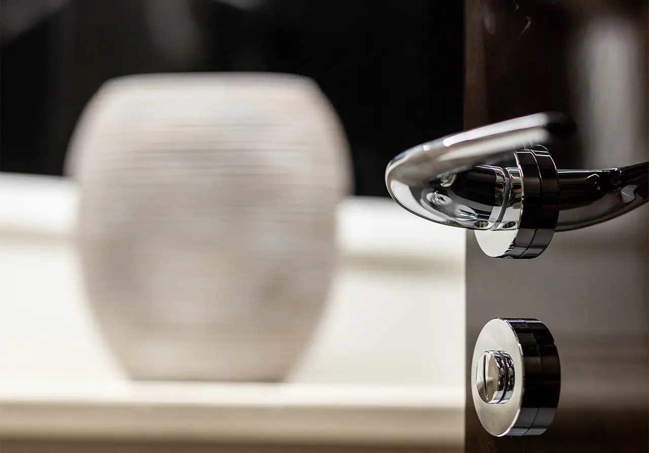 Close-up of a chrome door handle and lock on a wooden door, with a blurred decorative vase in the background.