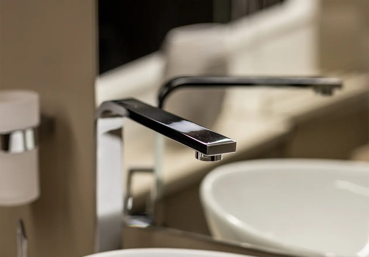 Close-up of a modern chrome tap over a white sink, showcasing a sleek design and the blurred reflection of a bathroom.