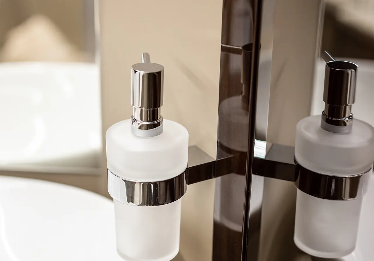 A sleek bathroom soap dispenser with a chrome pump, mounted beside a mirror and sink, reflecting a modern design.