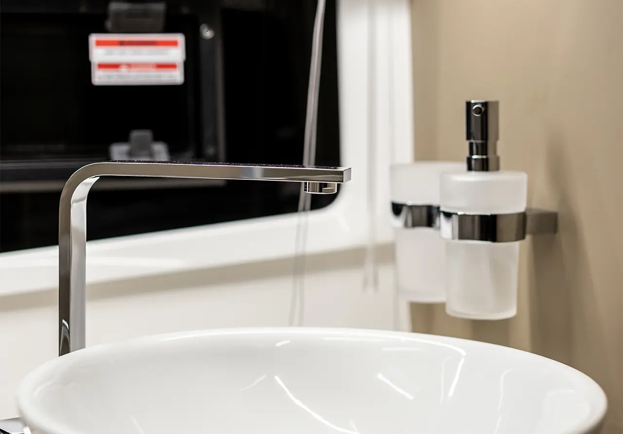 Modern bathroom sink with a sleek chrome tap, flowing water, and two frosted glass containers on a light wall.