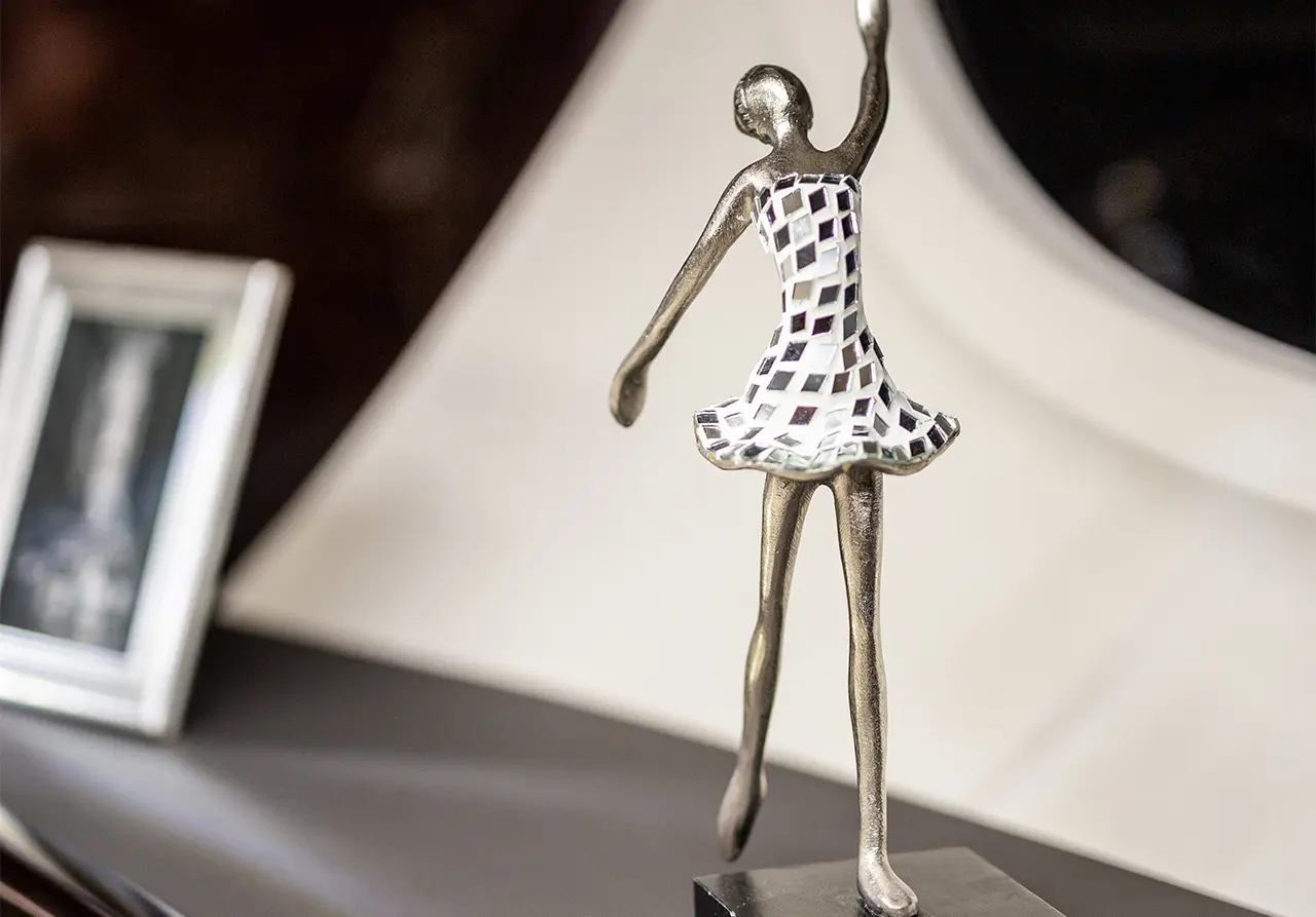 A metallic dancer figurine with a patterned dress raises one arm, displayed on a sleek surface near a photo frame.