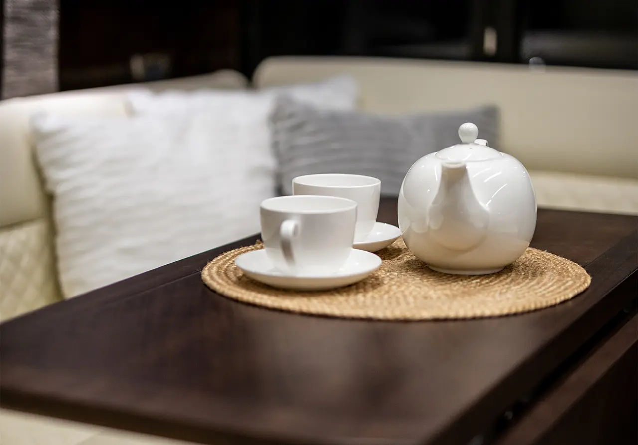 A white teapot and two cups on a wicker mat, set on a wooden table with soft cushions in the background, creating a cosy atmosphere.