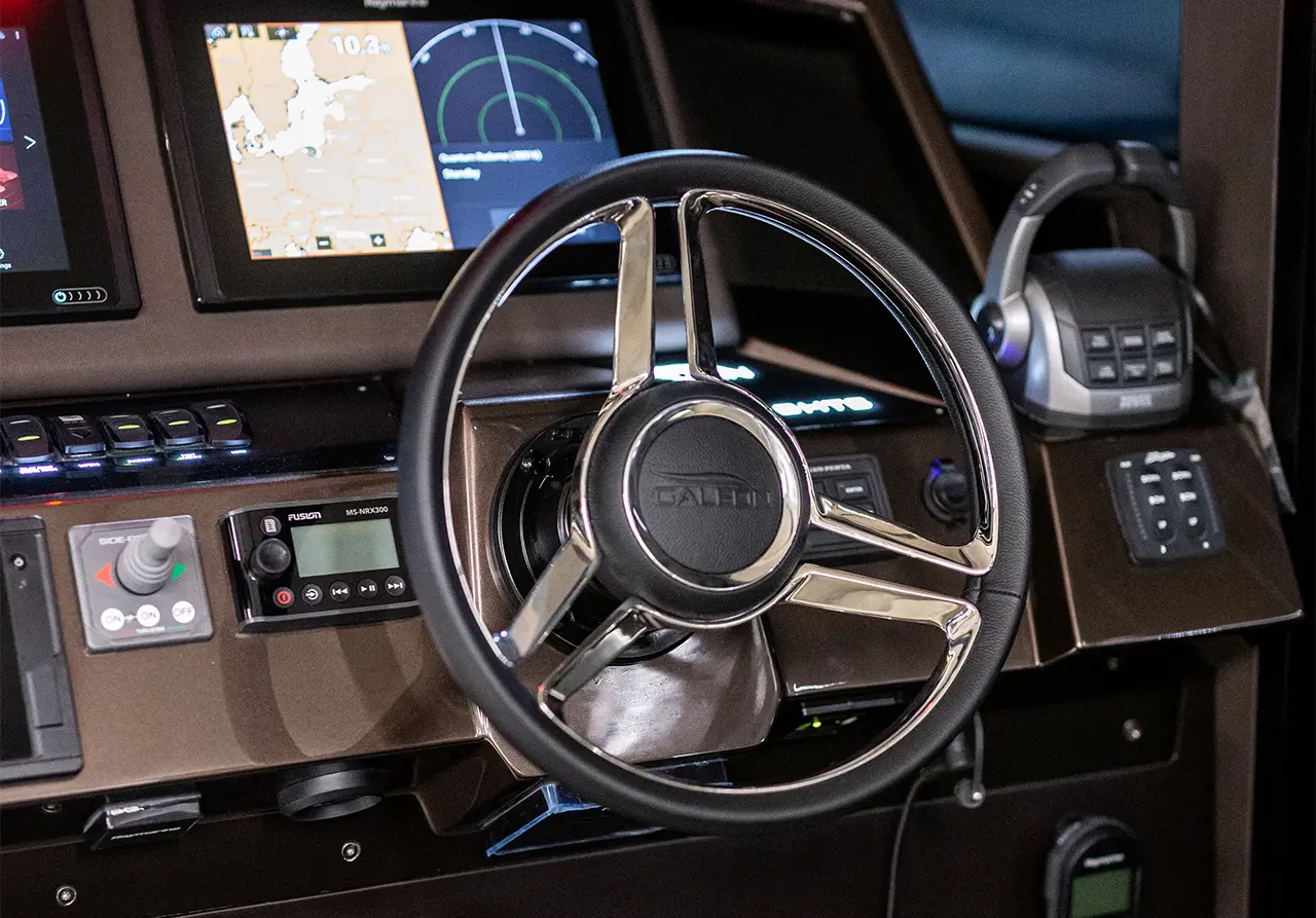 Boat steering wheel with navigation displays showing maps and radar, alongside various control buttons and audio device.