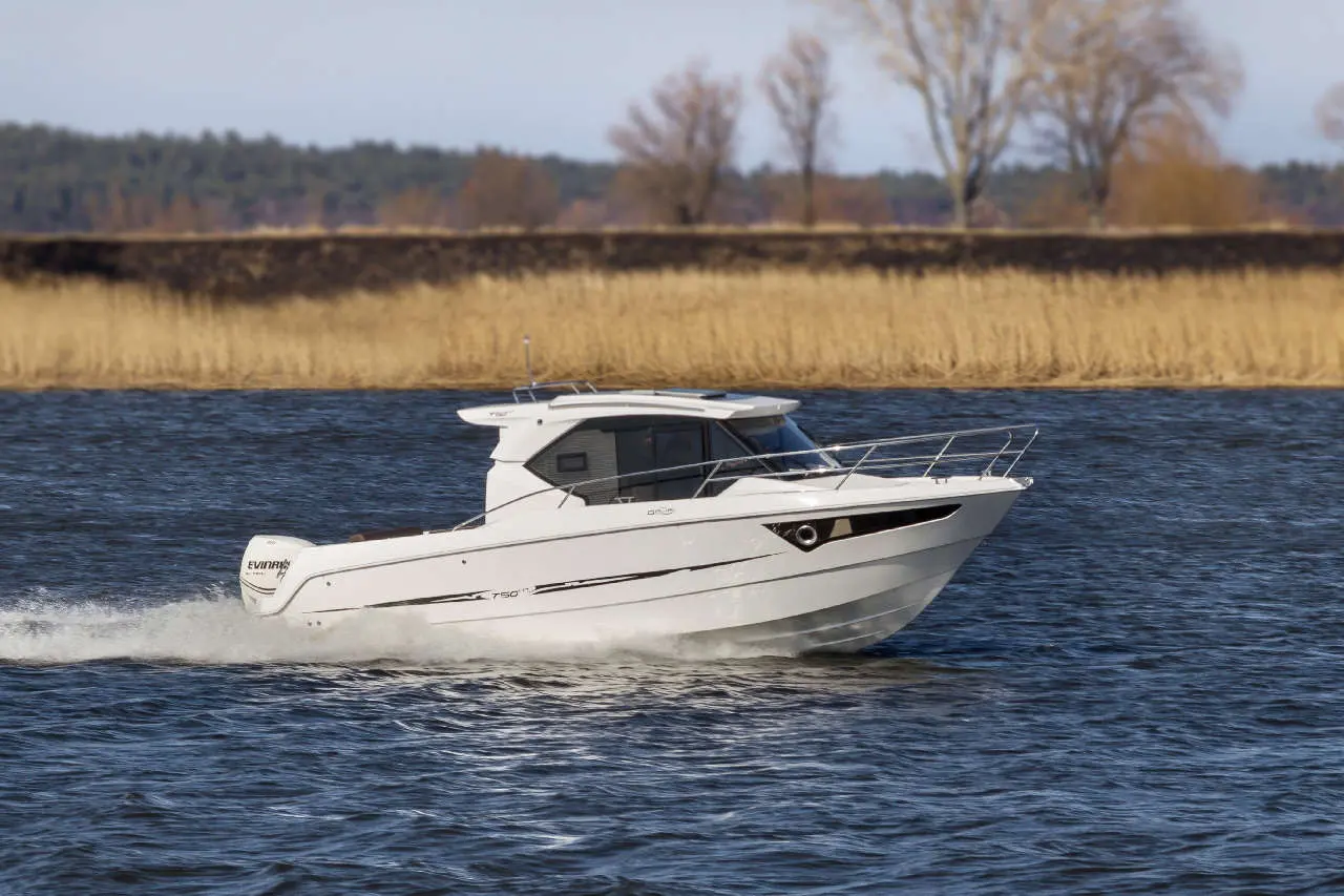 White powerboat navigating a calm river, with tall grass and trees in the background under a clear sky.