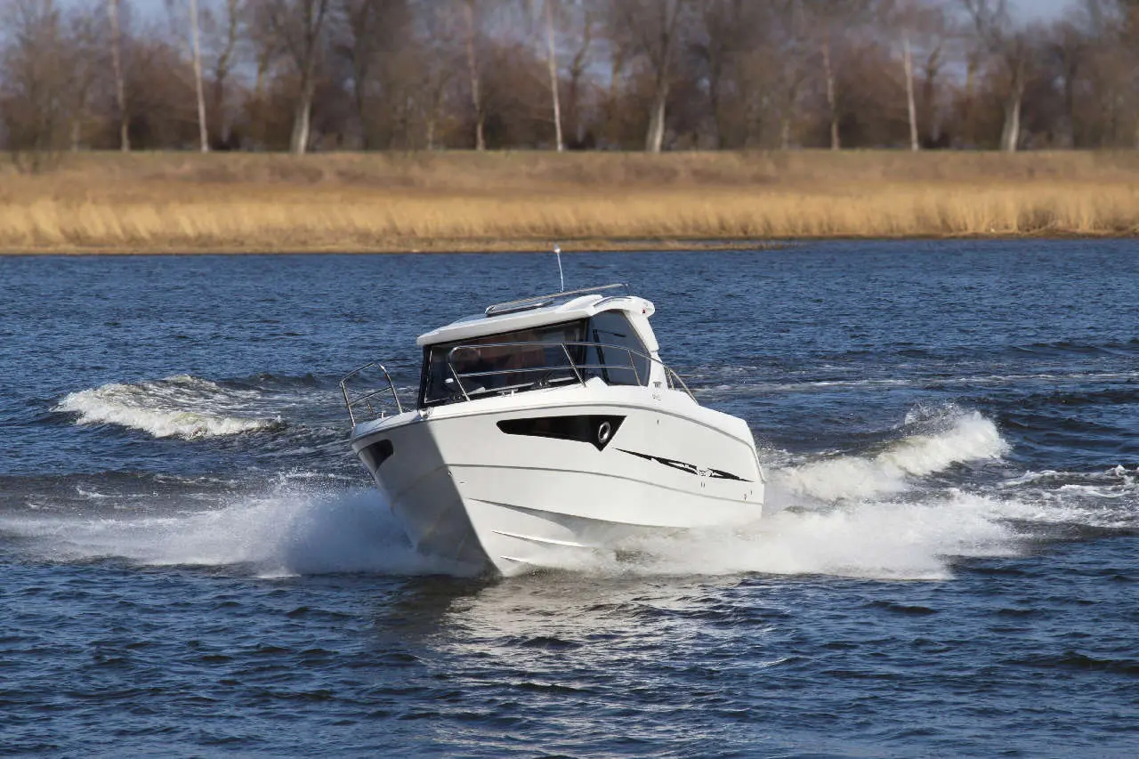 A white motorboat sails through a blue river, creating waves and splashes against a backdrop of trees and grass.