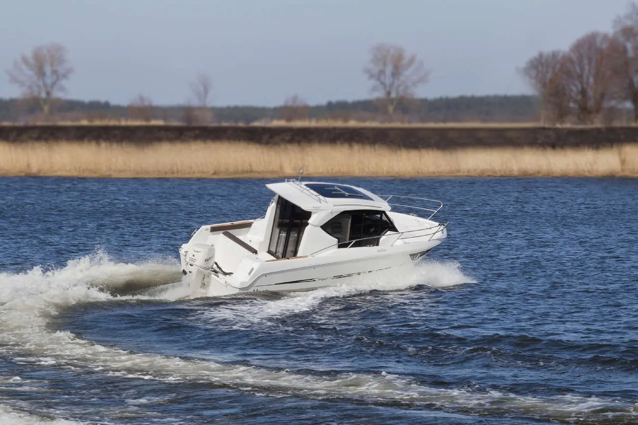 A white motorboat speeds across water, leaving a wake behind, with trees and dry grassland visible in the background.