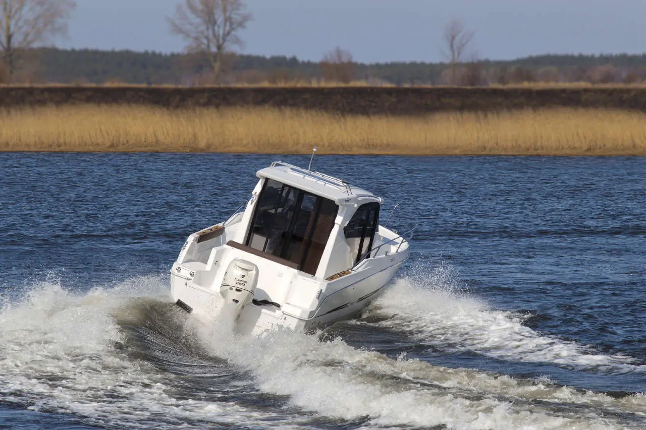 A white motorboat speeds through rippling water, creating waves, with grassy land and trees in the background.