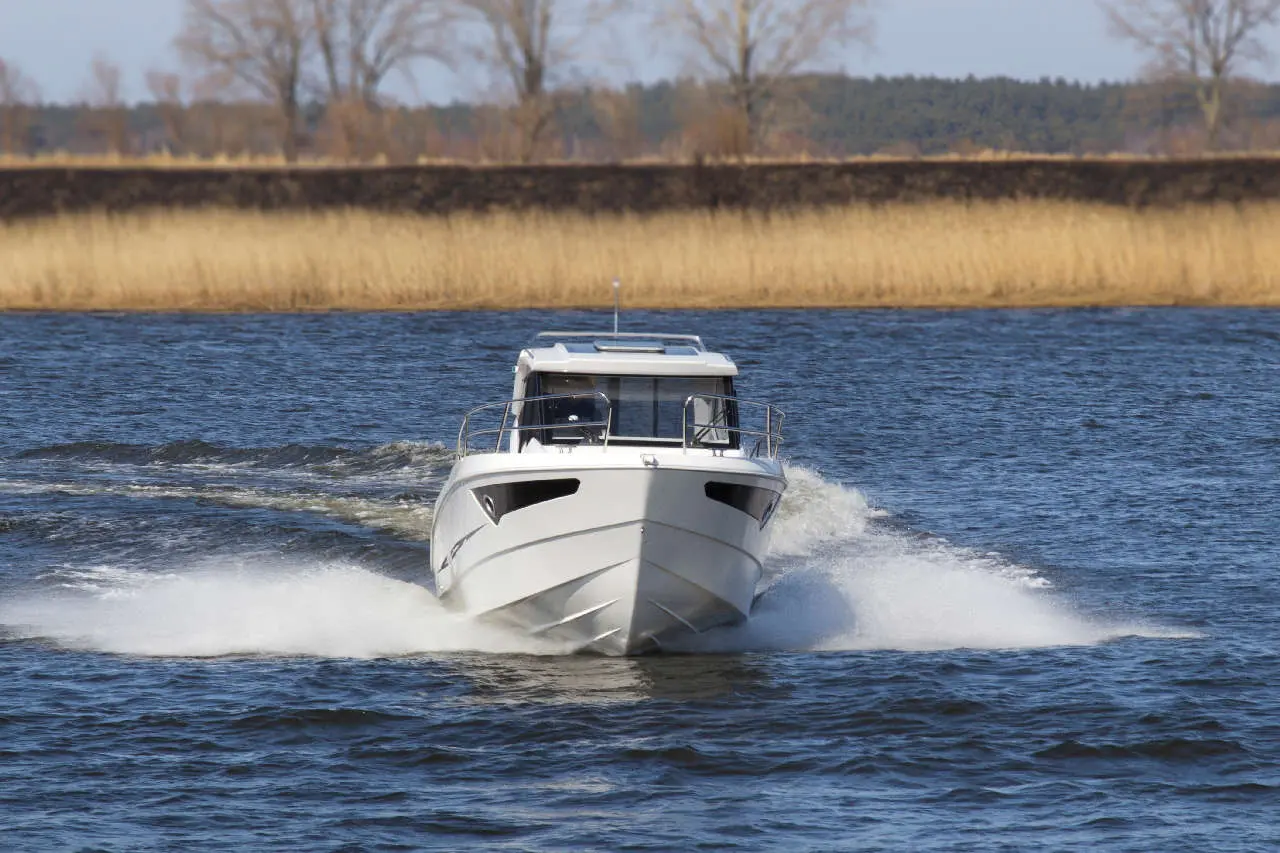 A white motorboat cuts through a calm river, creating a spray, with reeds and trees lining the water's edge.