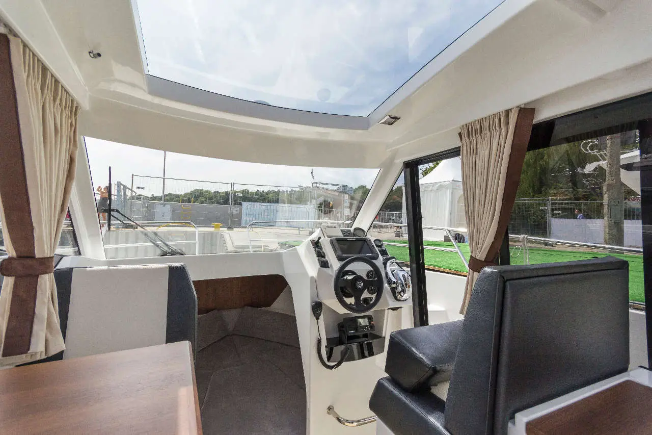 Interior view of a modern boat with a steering wheel, dashboard, and large windows, featuring sunlight and light curtains.