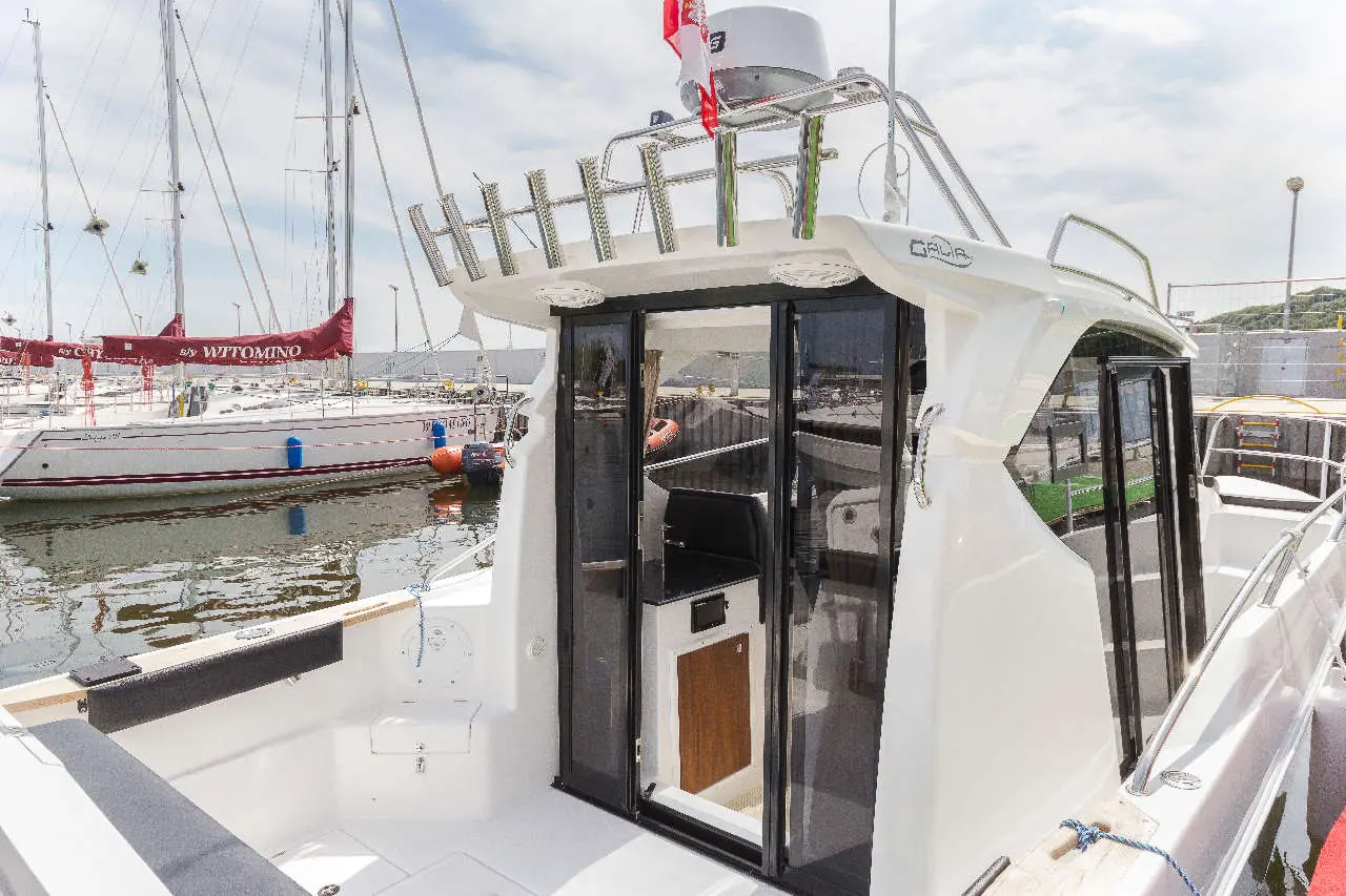 View of a boat with glass doors, moored near sailing vessels and a marina, featuring a red flag labelled 'WITOMINO'.