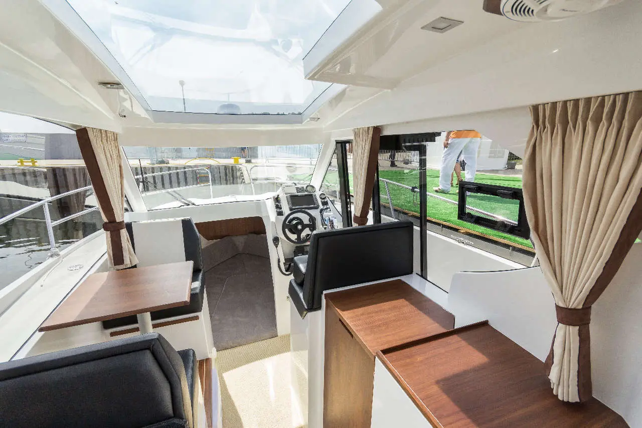 Interior of a modern boat featuring a steering wheel, seating area, and large windows, with a view of the harbour outside.