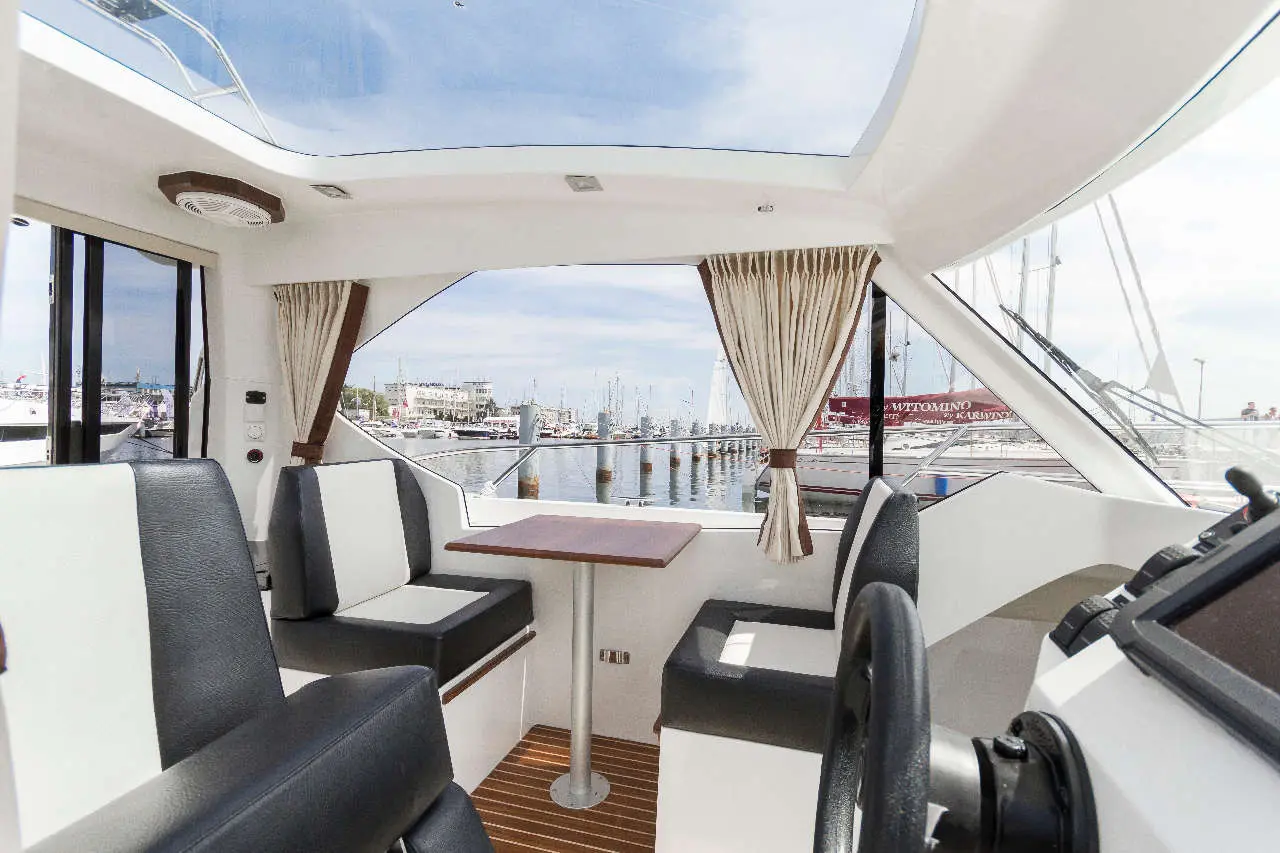 Modern boat interior with seating, table, and panoramic windows overlooking a marina filled with yachts and sailboats.