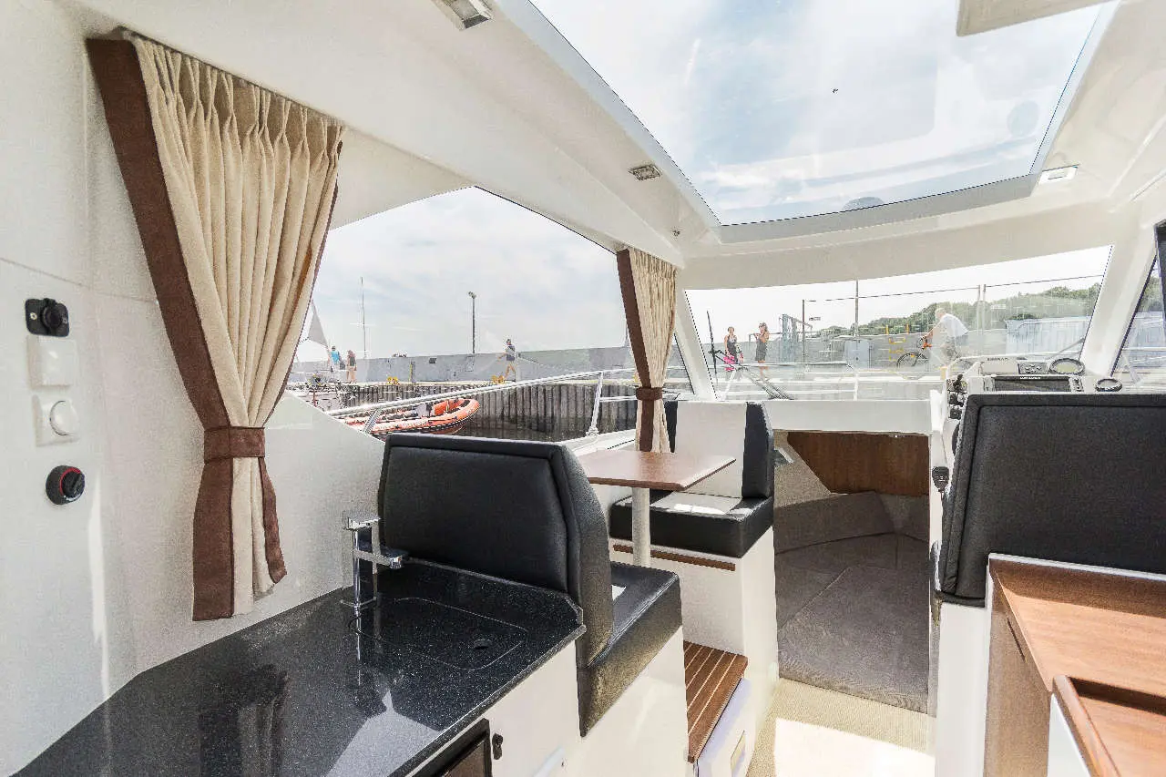 Interior of a modern boat featuring a kitchenette, dining area, and large windows overlooking a marina and people walking.