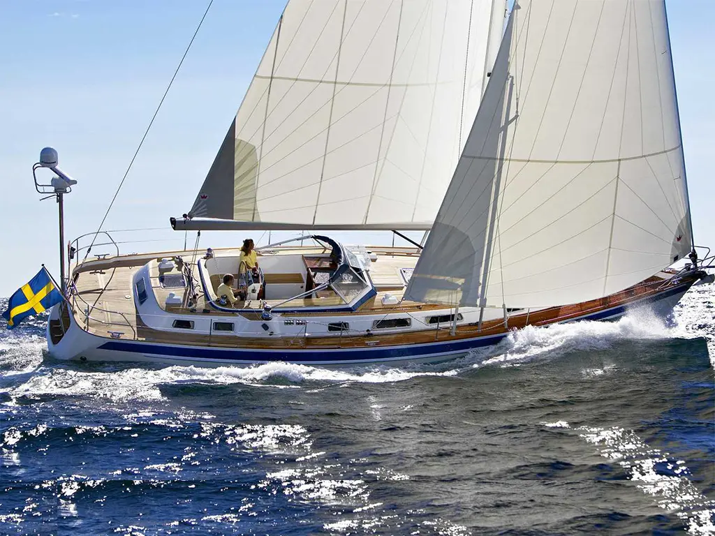 A sailing yacht with white sails and a Swedish flag, navigating through bright blue waters under a clear sky.