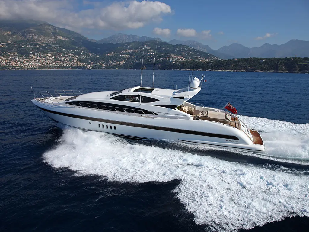 Luxury yacht cruising on calm waters, leaving a white wake, with mountains and a coastal town in the background.