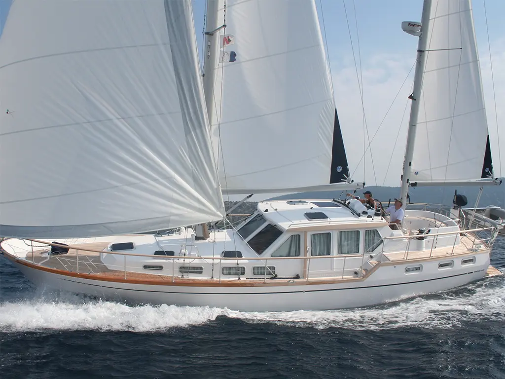 A white sailing yacht with a wooden deck glides through blue waters, featuring raised sails and two crew members on board.