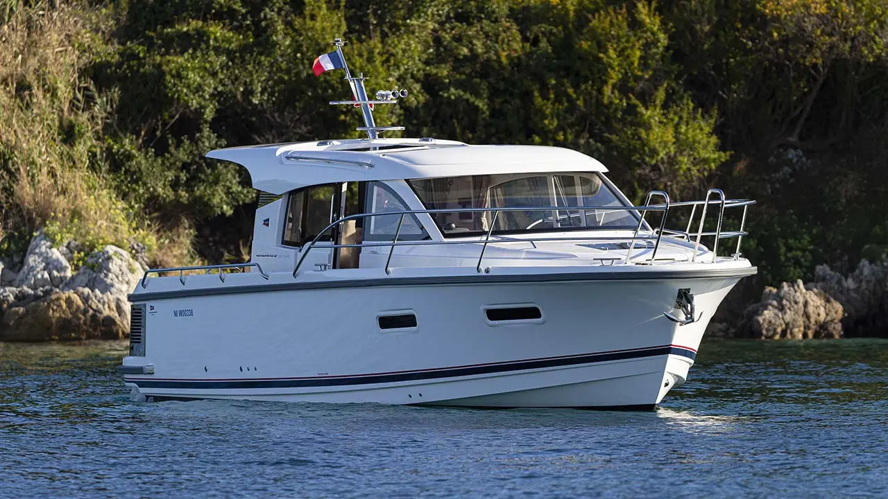 White cabin motorboat cruising on blue water beside a rocky, wooded shoreline, flying a French flag.