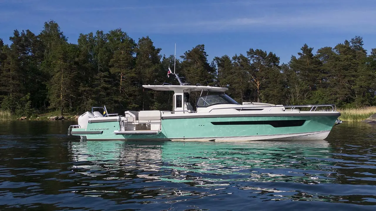 Turquoise cabin motorboat floating on calm water beside a forested shoreline under a clear blue sky.